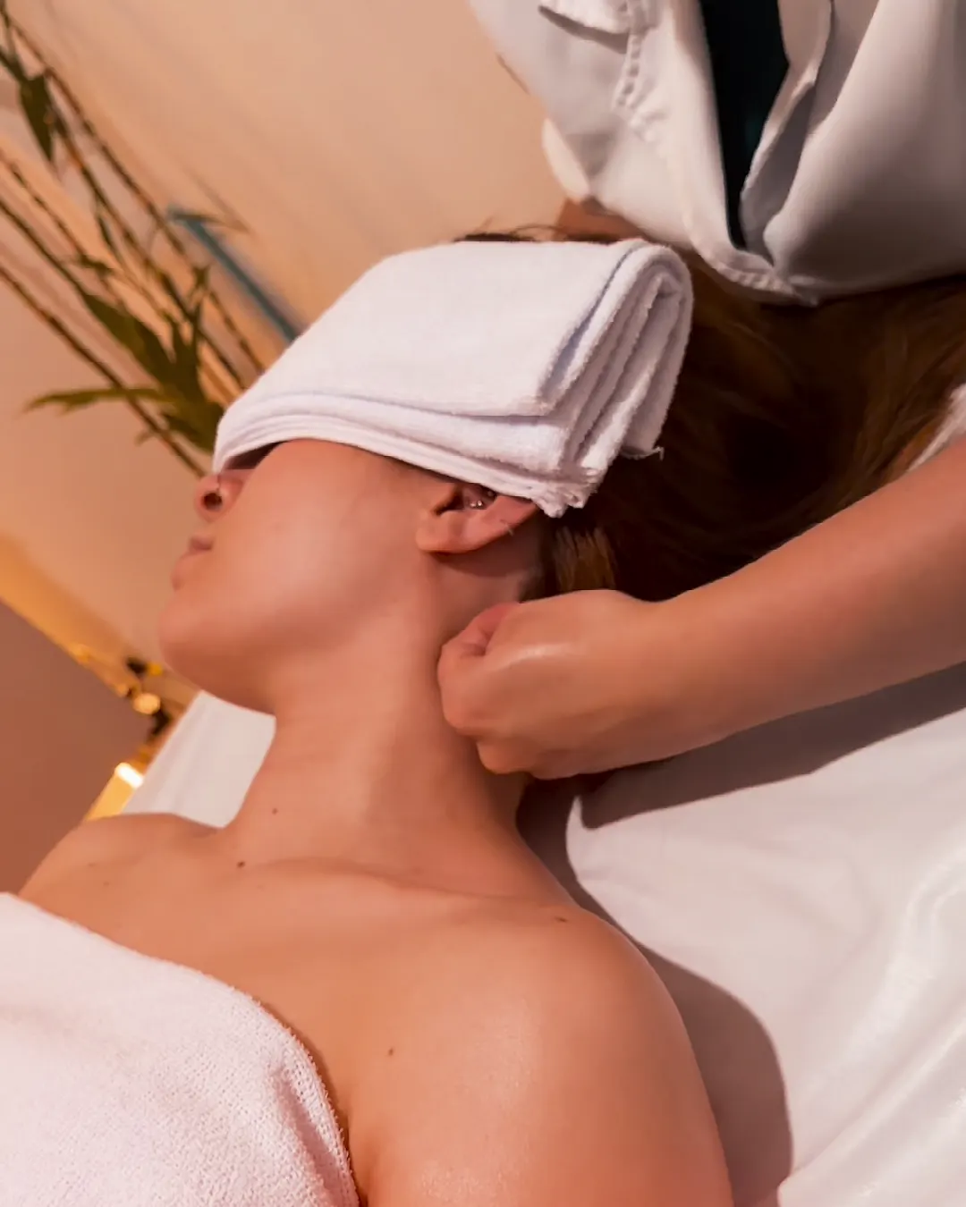 Massoterapeuta massageando o pescoço de cliente deitada na maca, com toalha sobre os olhos, no estúdio em Campo Comprido